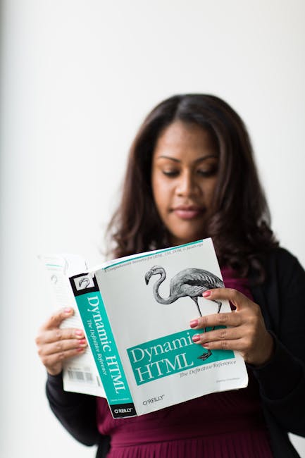 A woman with long dark hair, dressed in a dark blazer and maroon top, is standing indoors against a plain white wall. She is holding an open technical book titled 'Dynamic HTML,' which features a larg